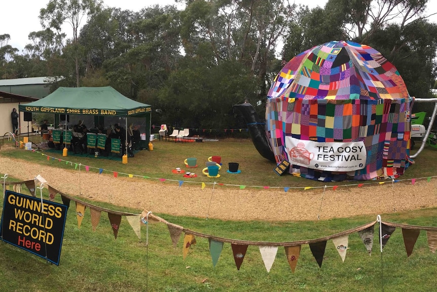 Giant teapot structure draped with colourful knitted squares sitting in a field