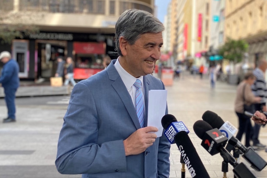 A man wearing a suit holds a piece of paper in front of him in front of shops