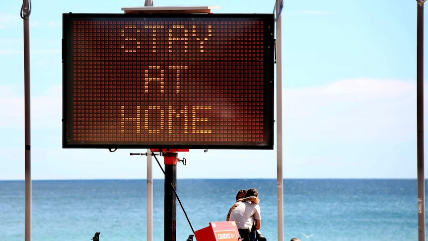 a sign saying stay at home on the beach where two people are hugging