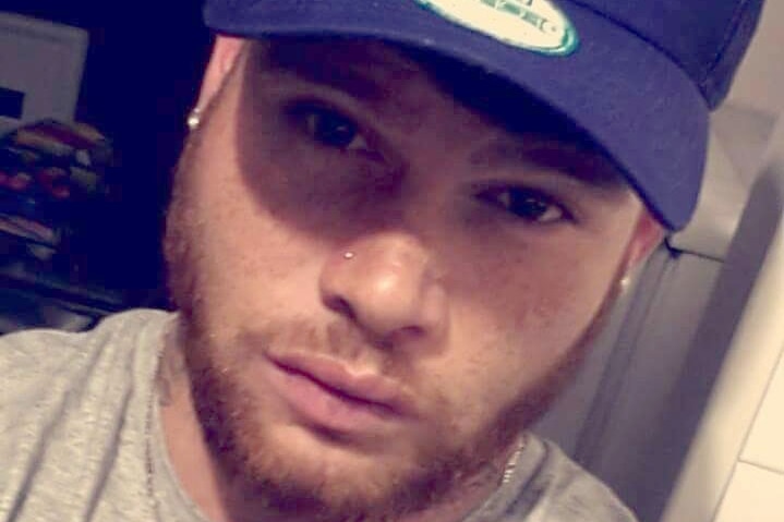 A young man with a short orange beard and earrings stares into the camera wearing a baseball cap.