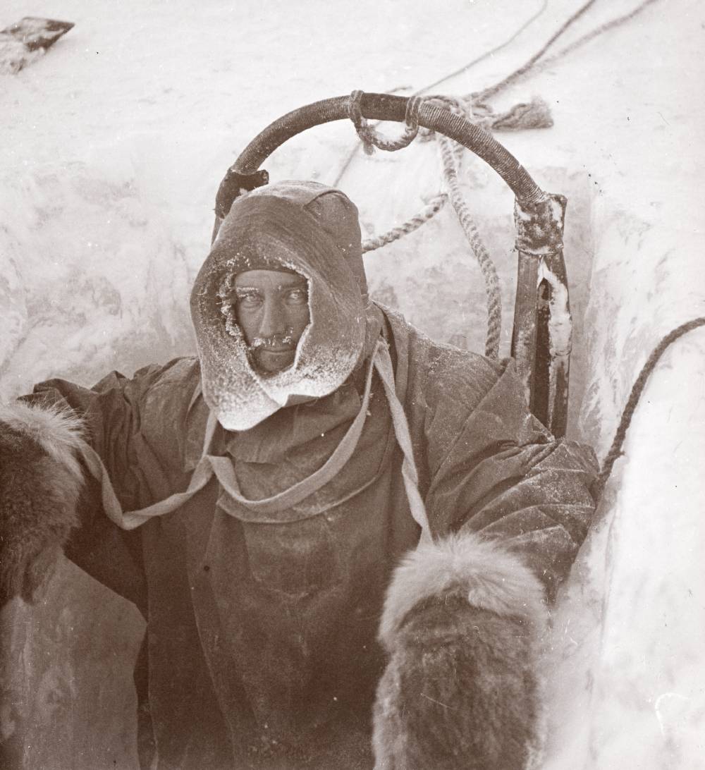 A man dressed in heavy, thick clothing looks up at the camera. He is standing in a hole in the ice.