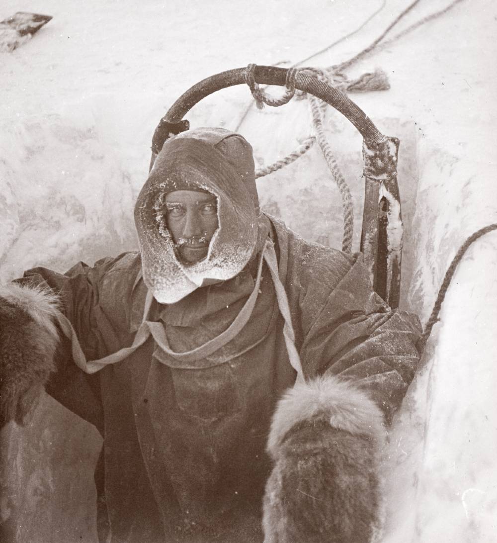 A man dressed in heavy, thick clothing looks up at the camera. He is standing in a hole in the ice.