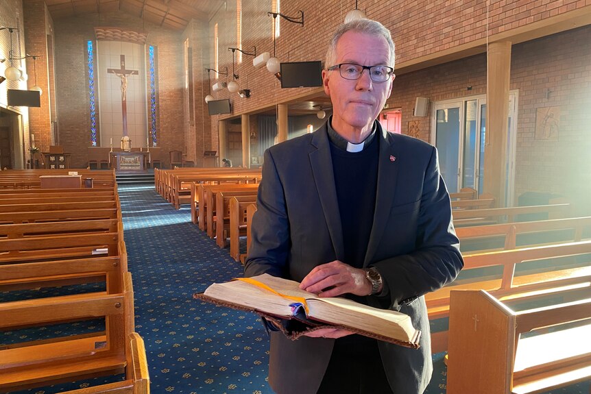 A man in priest's clothing stands in church holding an open bible as he looks at the camera.