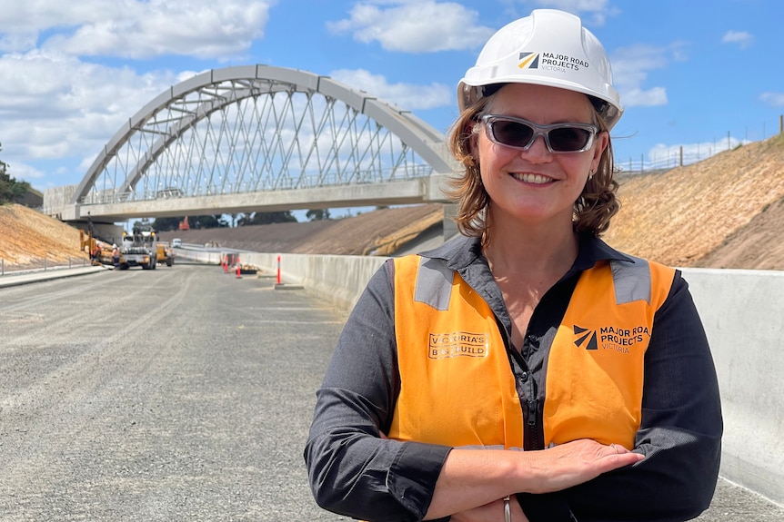 Allison is in a high-vis vest, glasses and hard hat standing on a gravel road in front of the new Kilmany rail bridge. 