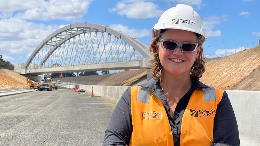 A woman in a high-vis vest, sunglasses and hard hat standing on a gravel road in front of an arc-shaped rail bridge.