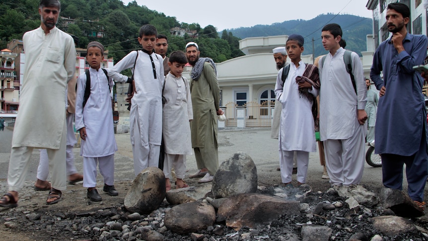 A group of Pakistani men and boys stand around a pile of burnt rocks and wood in a town square.