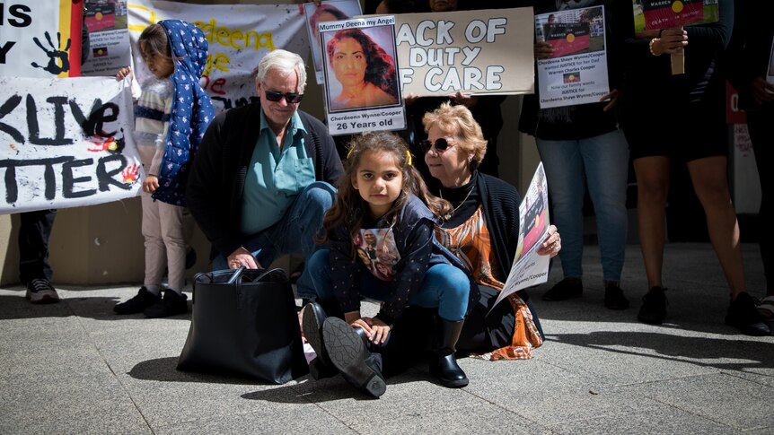 A girl crouches in front of two adults, surrounded by people holding signs.