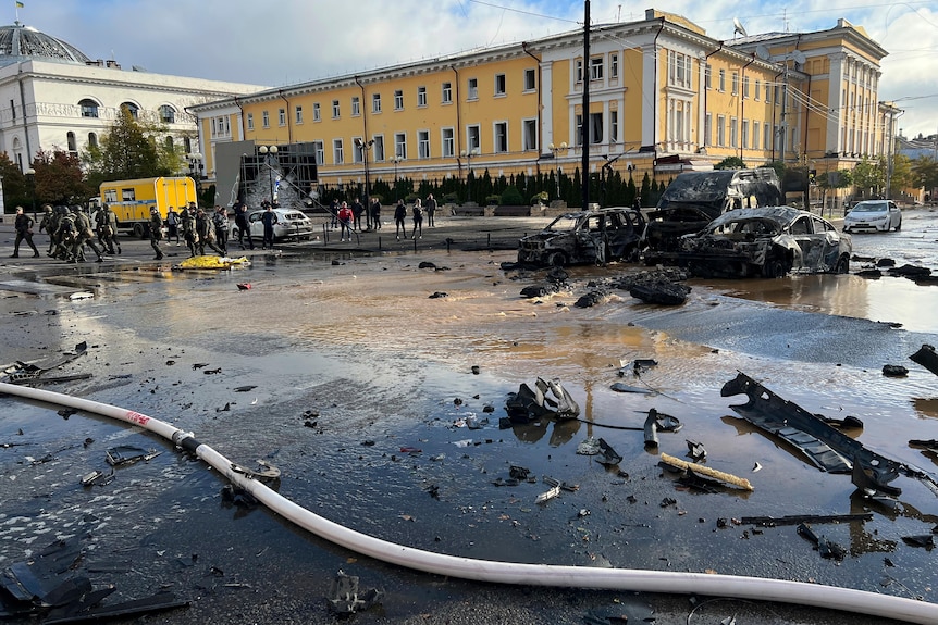 Wrecked, burnt cars sit in the middle of a destroyed, partially flooded road surrounded by debris.