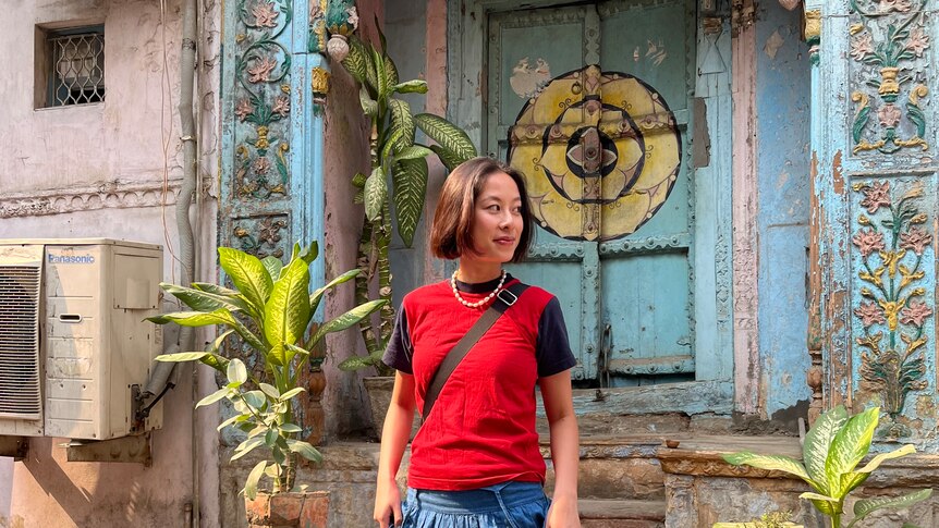 Write Maggie Zhou stands on steps in Dehli wearing a red tank over a black t-shirt and a long denim skirt.