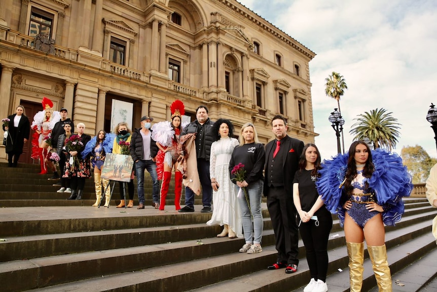 Men and women in colourful costumes stand in front of the steps of an old building. 