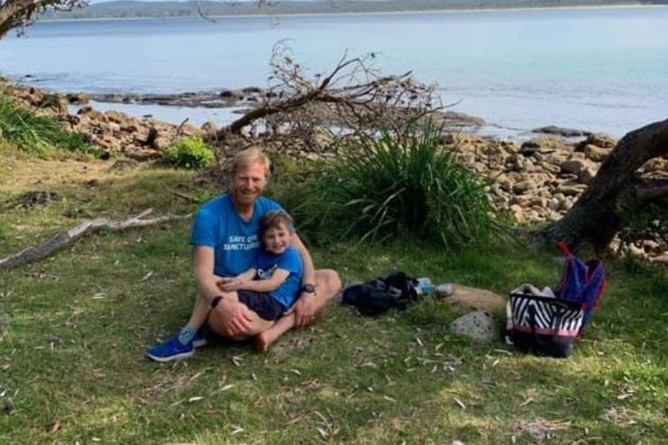A smiling blond man, with a smiling boy on lap, sit on the grassy foreshore near a beach, couple of bags near them.