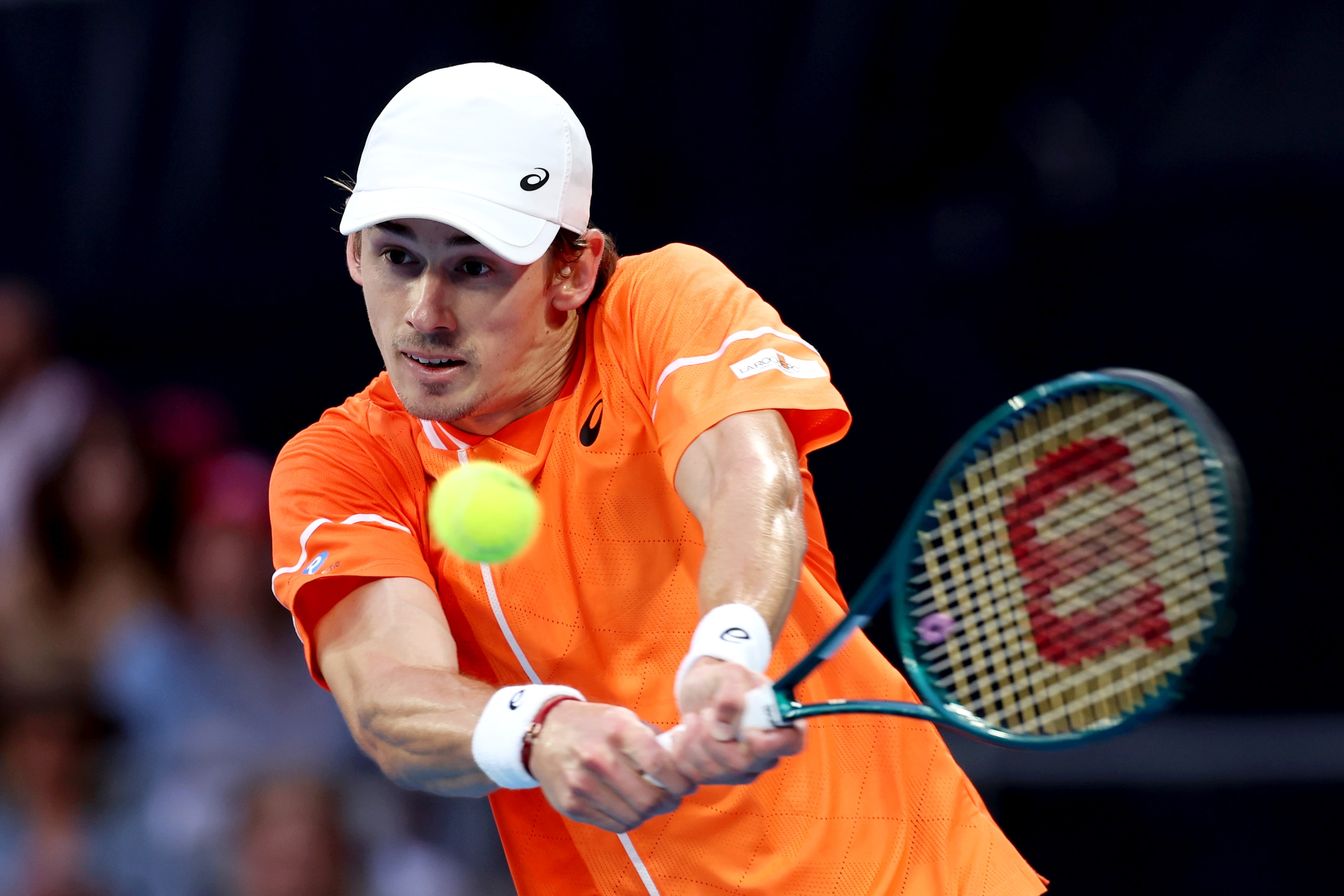 A male tennis player, an orange shirt, white hat, plays a two-handed backhand.