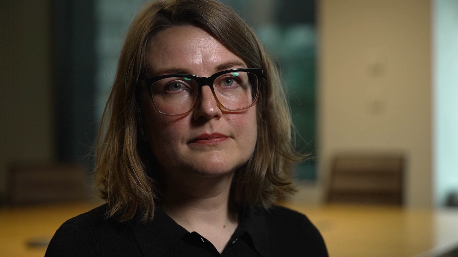 A woman with a brunette bob and glasses sits in a conference room.