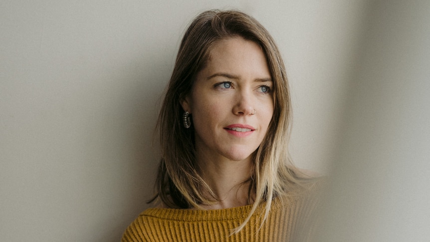 A young blonde woman is leaning against a blank wall with her arms folded, looking into the distance. Ausnew Home Care, NDIS registered provider, My Aged Care