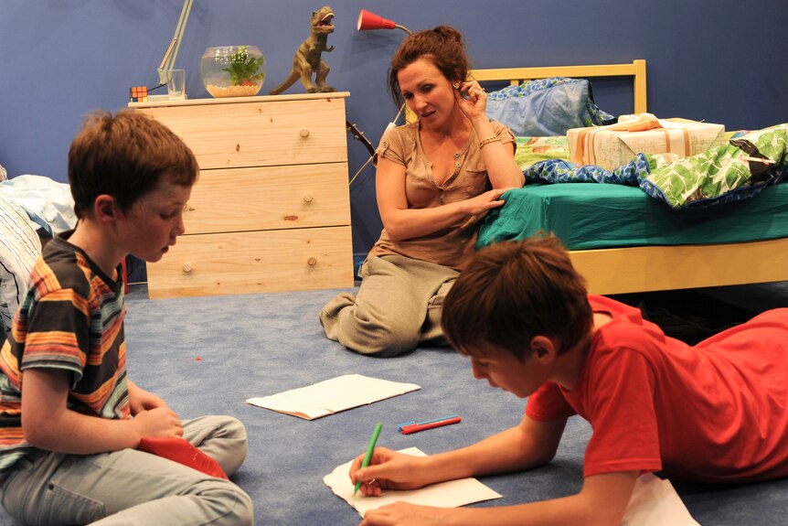 In a kids' room, a mother sits leaned against a bedframe, as two boys draw with textas and paper on the floor