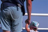 A father holds his toddler daughter's hand while they both stand on a pier.