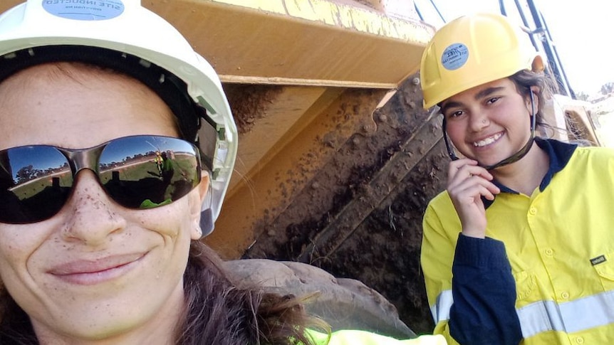 Two women in high vis wearing hard helmets stand next to a tractor.