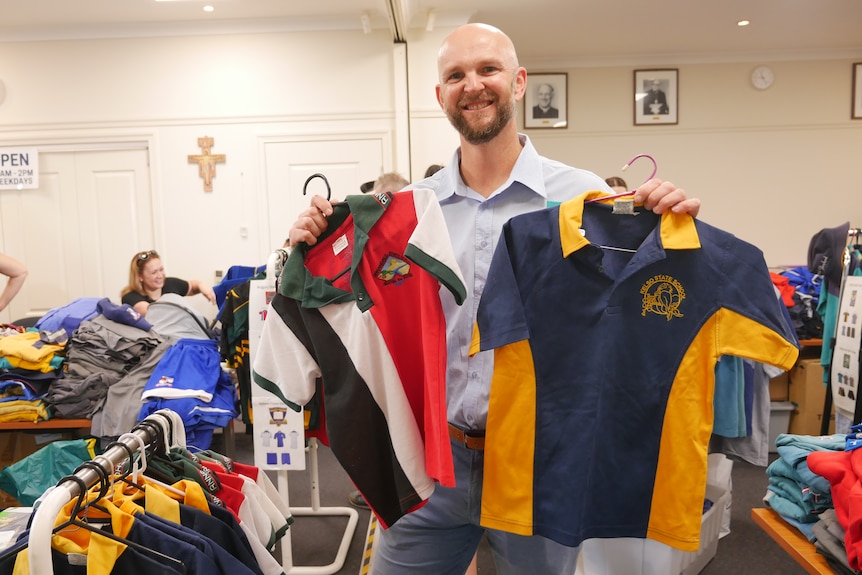 a bald man stands in a room holding two polo shirts, smiling
