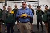 A man smiles while holding a volleyball and standing in the middle of a group of volleyball players.