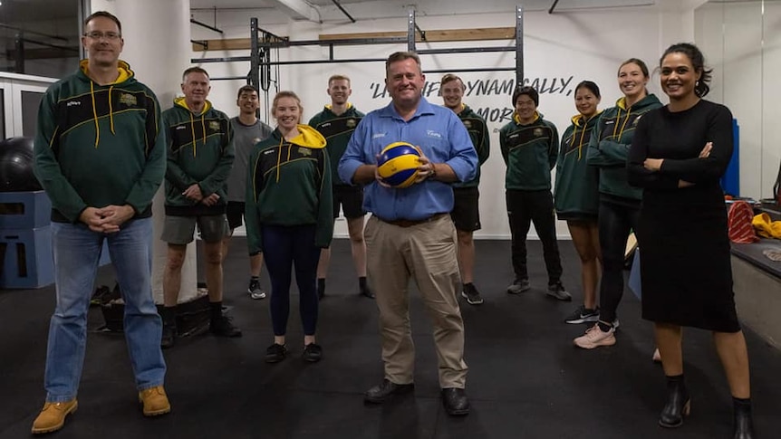 A man smiles while holding a volleyball and standing in the middle of a group of volleyball players.