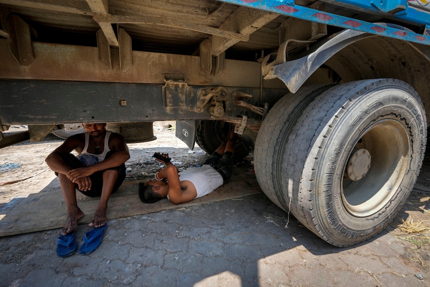 Two men in white singlets sit or lie under the chassis of a truck.