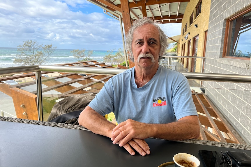 Aboriginal man with Aboriginal flag on a blue T-shirt on a balcony with the beach behind him. 