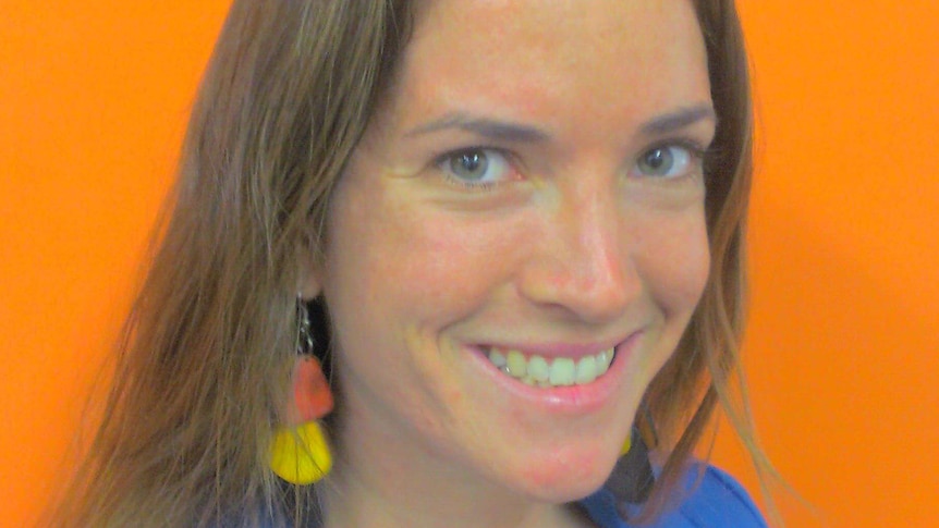A close-up of a woman with shoulder-length brown hair, with a blue jacket, in front of an orange background.