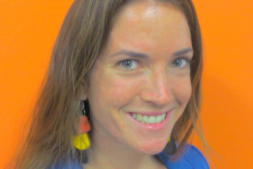 A close-up of a woman with shoulder-length brown hair, with a blue jacket, in front of an orange background.