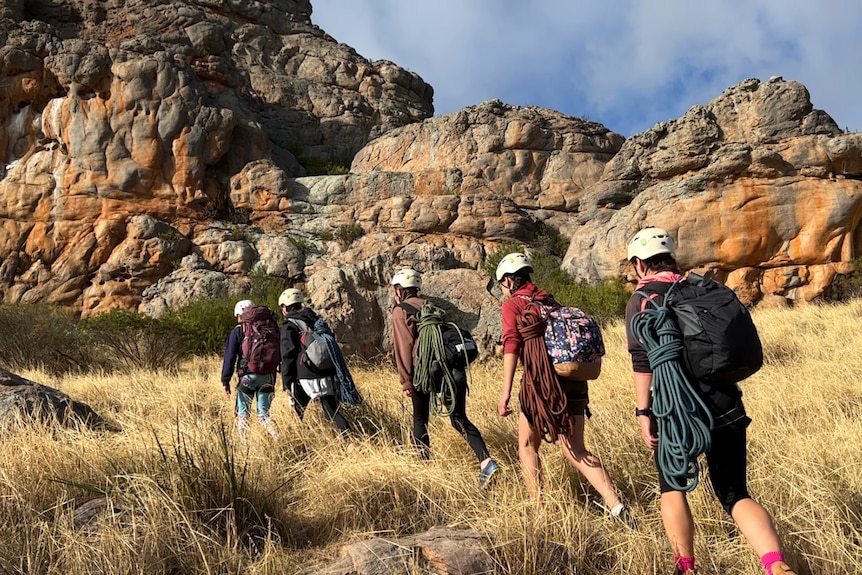 girls and women walk single file among dried grass and rock towards the base of an orange sandstone mountain carrying backpacks 