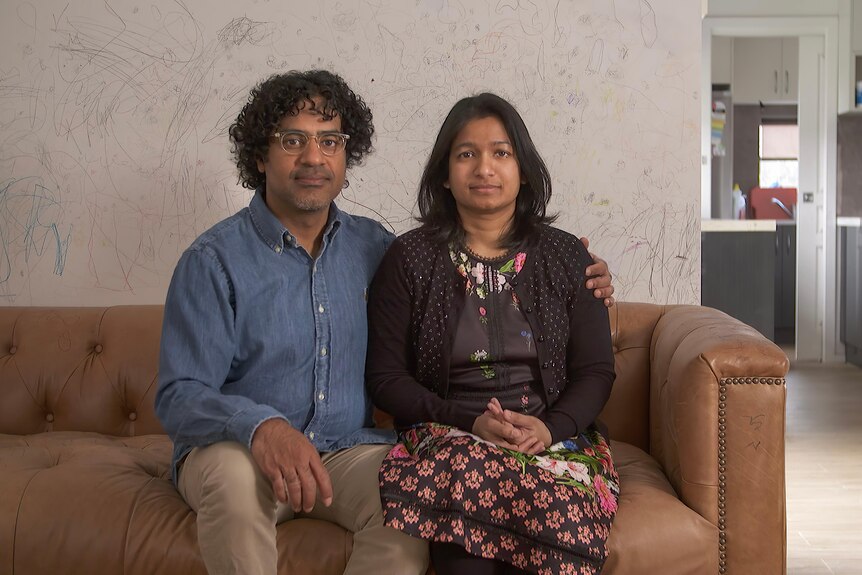 A couple sit on a couch looking directly at the camera with serious expressions
