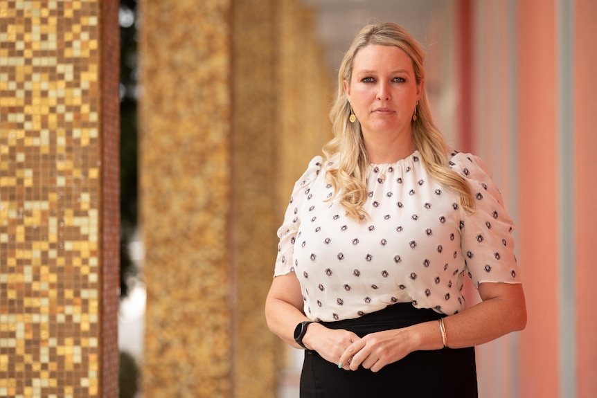 A blonde woman wearing a spotted shirt stands in front of a building