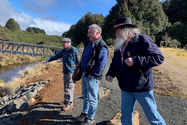 Three men stand at the edge of a dam, one pointing at it.