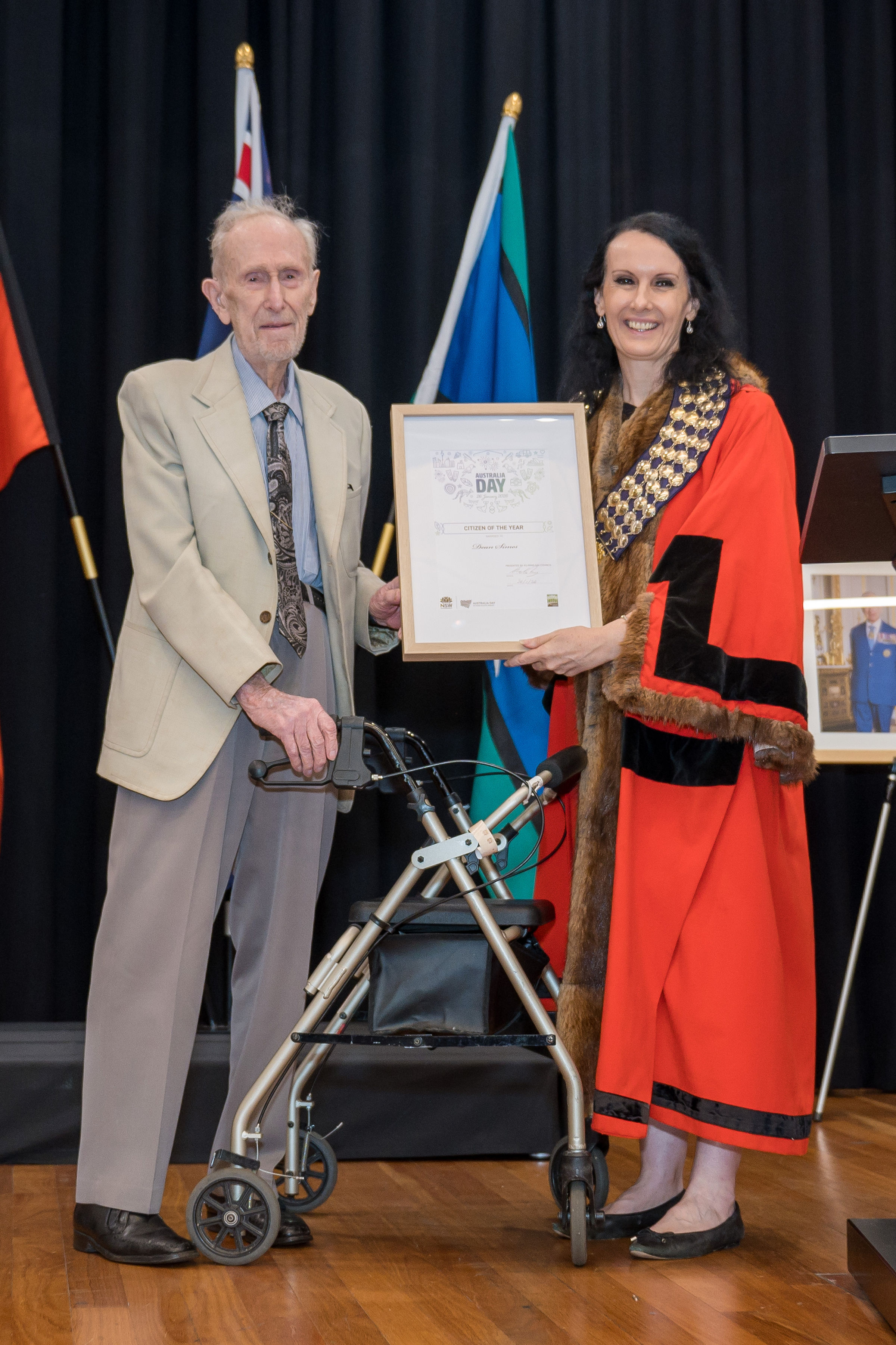 Un anciano sostiene un premio enmarcado junto a una mujer vestida con una túnica roja.