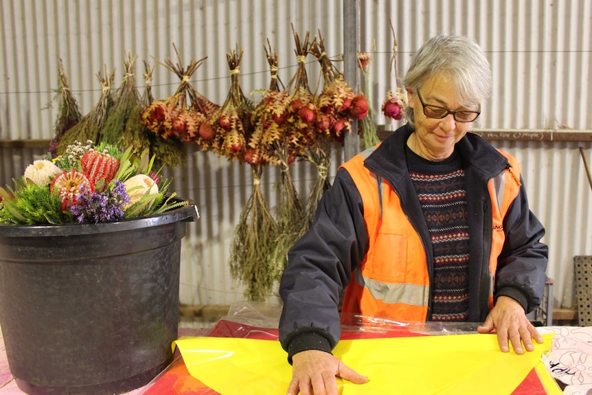 A woman smooths wrapping paper by a bucket of native flowers; dried bouquets hang on the shed wall.
