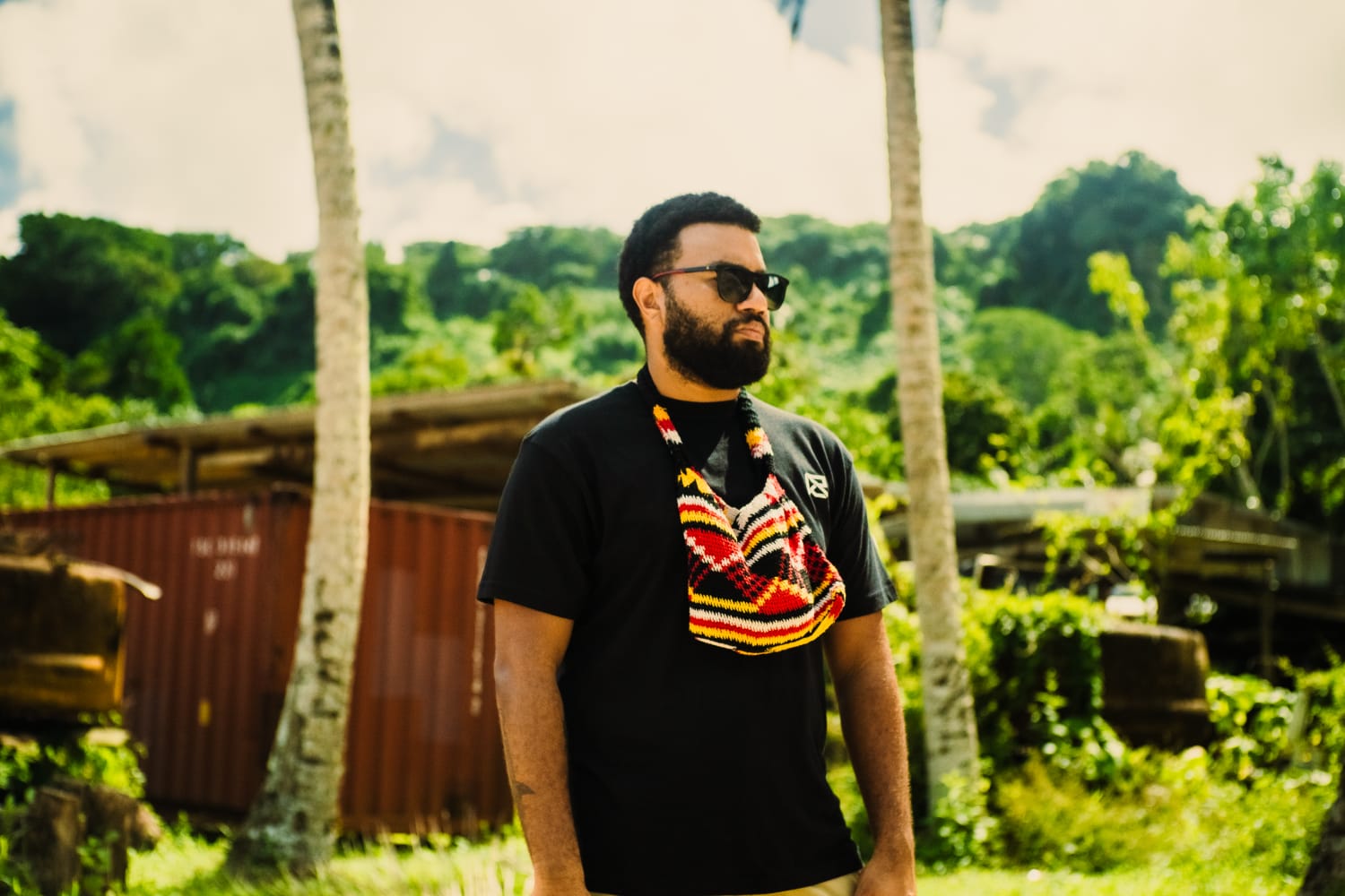 Profile mid view of artist Mantis from Vanuatu, green hills behind him. Wears red bilum around his neck and black sunglasses. 