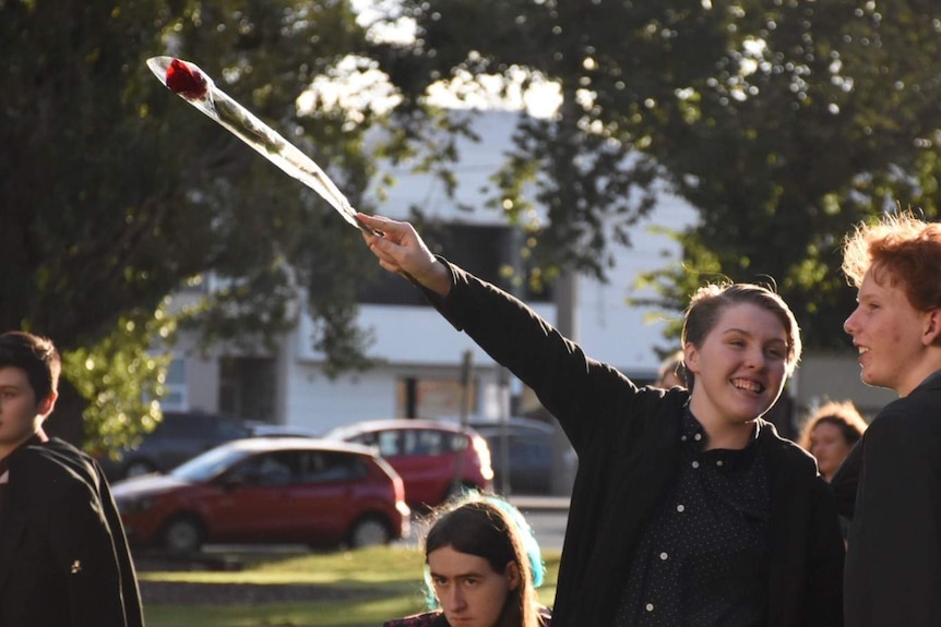 A young person holds a single rose and points it with their arm outstretched.