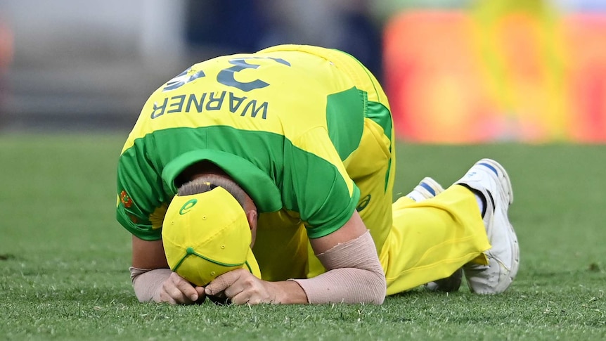 An Australian cricketer kneels with his head on the grass after injuring himself while fielding.