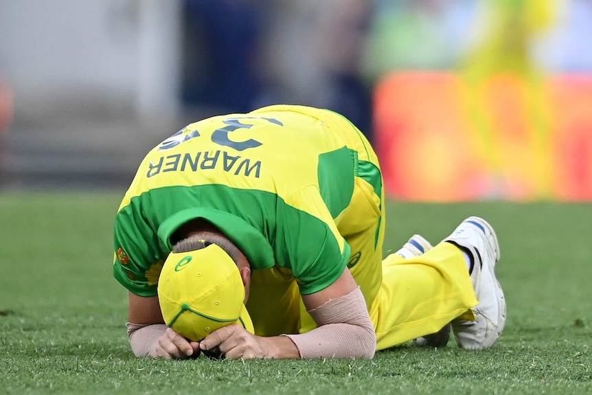 An Australian cricketer kneels with his head on the grass after injuring himself while fielding.