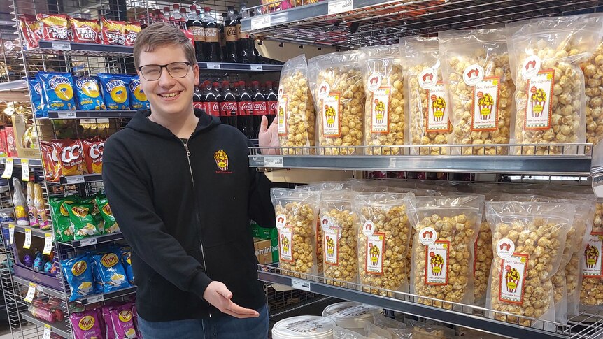 Sam stands smiling to the side of a shop end-of-aisle display of his popcorn.