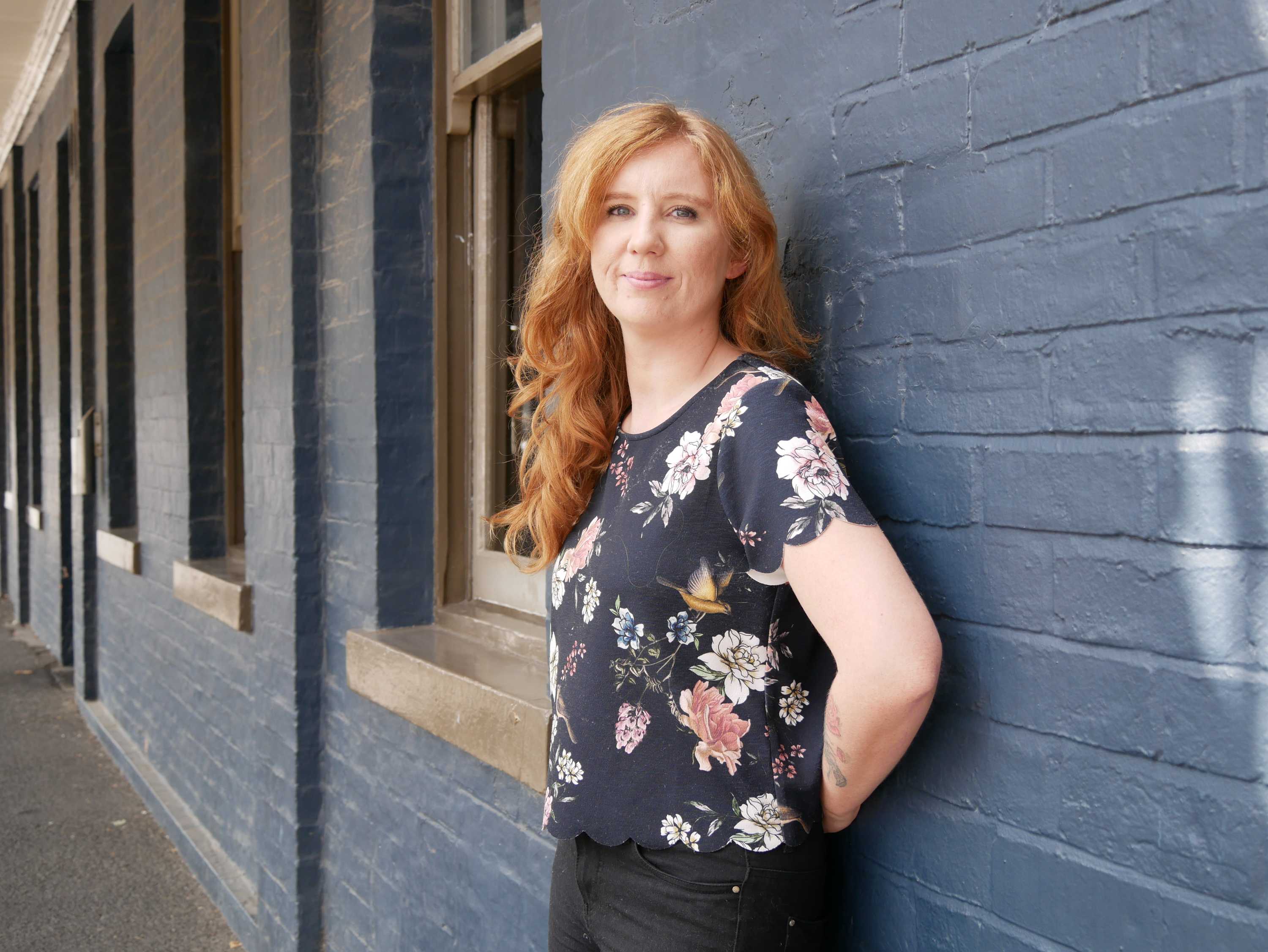 A woman with long red hair, standing in front of a blue brick wall, smiles to camera.
