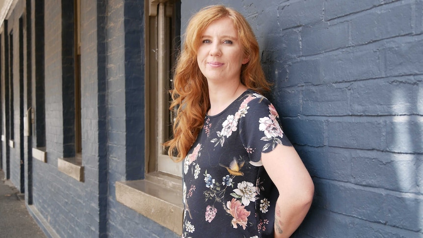 A woman with long red hair, standing in front of a blue brick wall, smiles to camera.