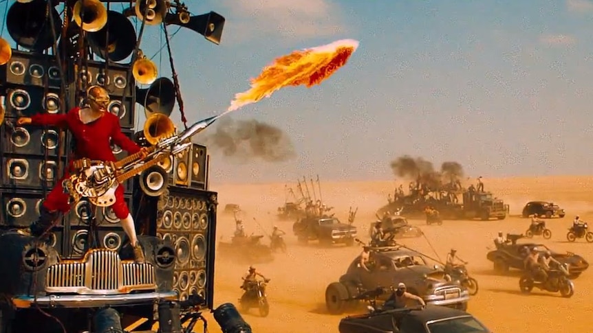 A man playing a flame-throwing electric guitar is suspended in front of a stack of speakers on a vehicle in the desert