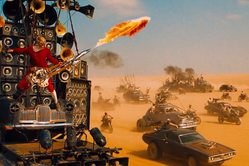 A man playing a flame-throwing electric guitar is suspended in front of a stack of speakers on a vehicle in the desert