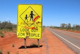 A road sign with graffiti warning to "look out for people" stands in a desert landscape.
