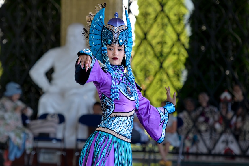 A performer dressed in bright blues and purples has her arms reached out during a dance. 