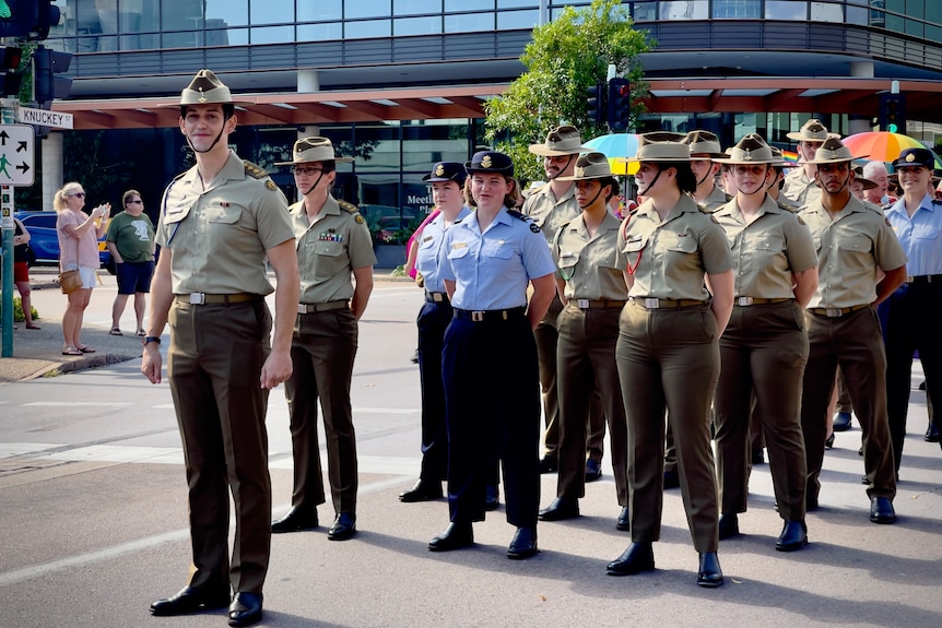 A smiling person wears female army uniform of tailored pants., shirt, hat. Leads a contingent of others, glass building behind.