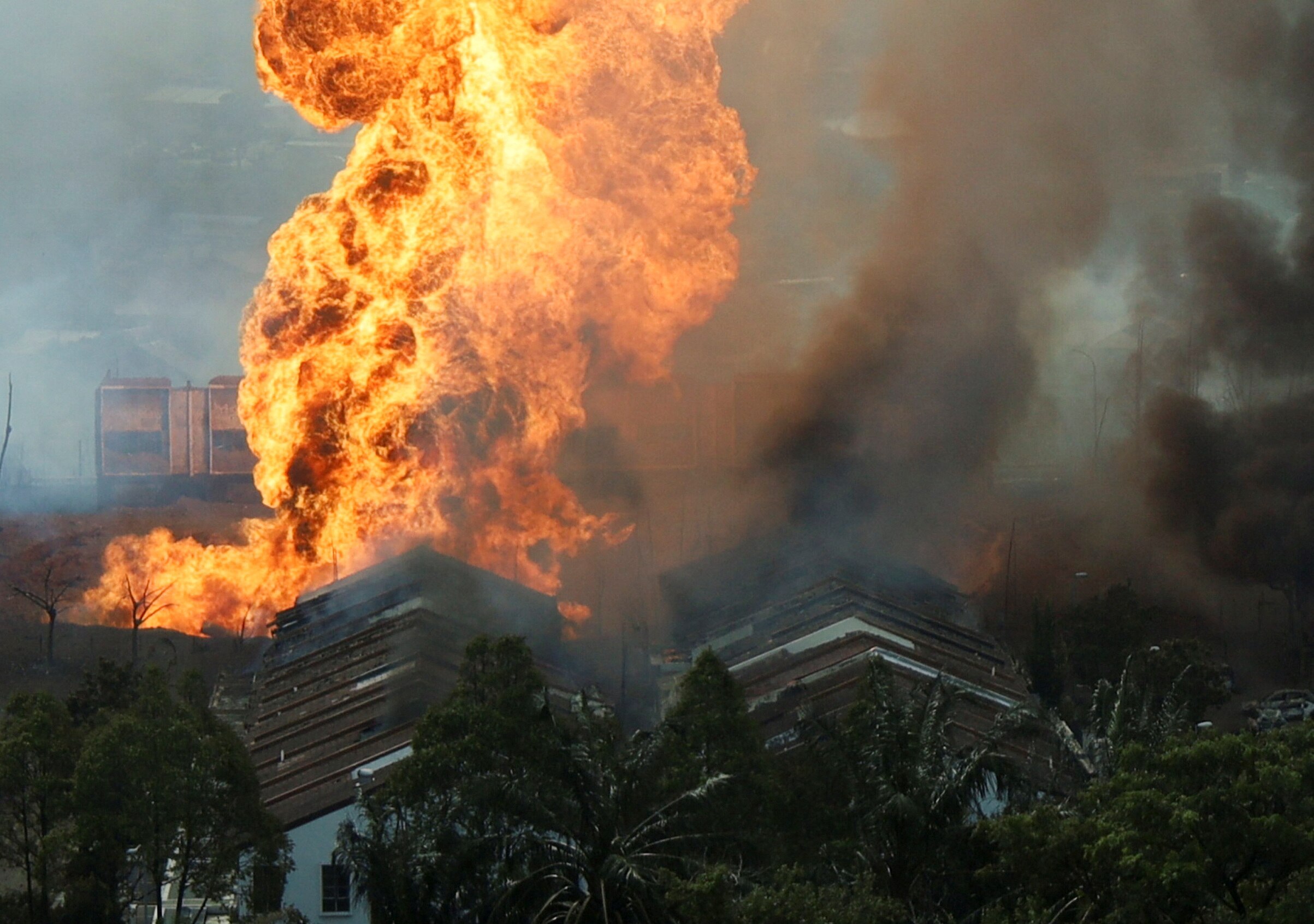A large, bright orange column of fire erupting from the peaks of brown house roofs surrounded by thick dark smoke plumes