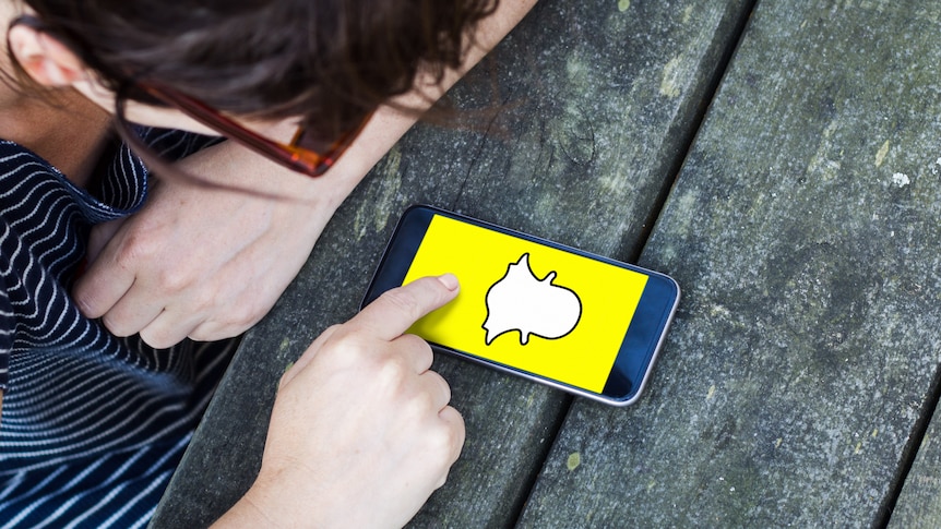 A teenager looks down at a smartphone screen displaying the Snapchat logo, a yellow screen with a white ghost cartoon