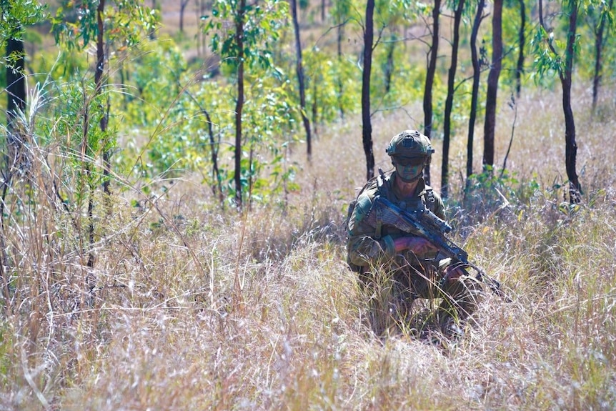 An Australian soldier kneels in thick scrub with camouflage paint on his face and his rifle at the ready.
