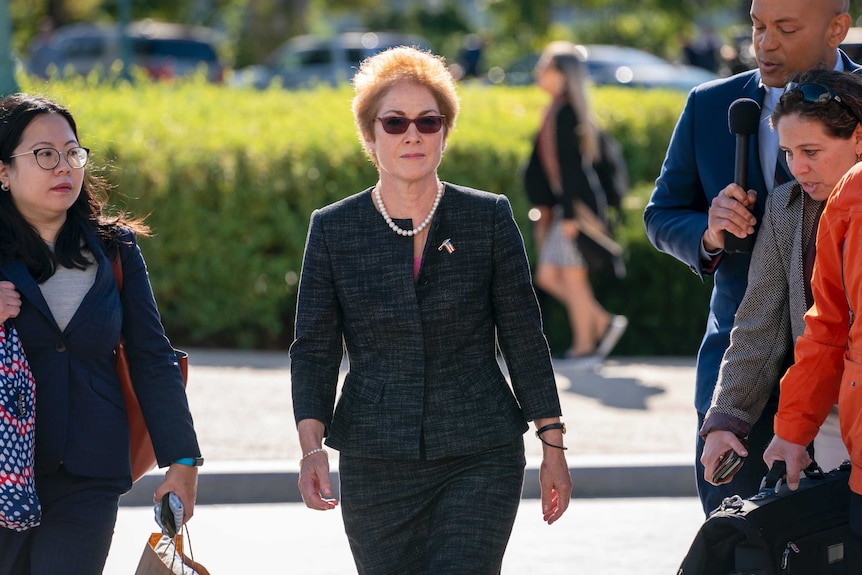 Marie Yovanovitch walks towards the camera with  stern expression. She wears sunglasses, a charcoal suit and a US flag brooch.